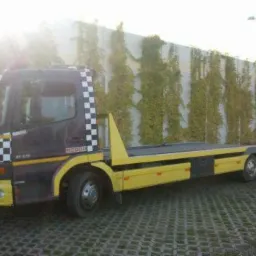 Żółto-czarna laweta z szachownicą na kabinie, stojąca na kostce brukowej przed betonowym murem porośniętym bluszczem.