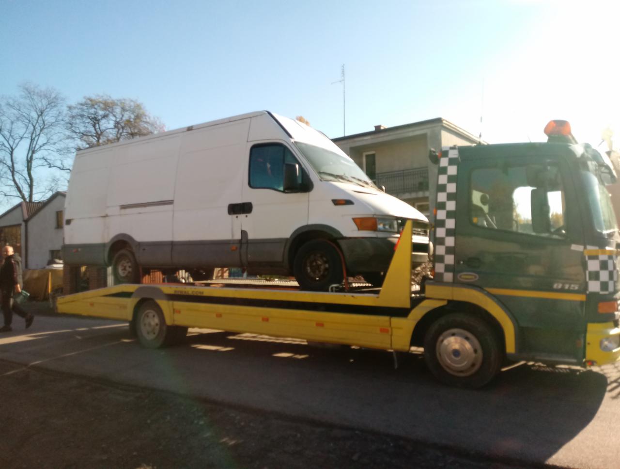 Biały bus na żółtej lawecie transportowany przez zielono-czarną ciężarówkę z szachownicą, widok z boku na tle zabudowań.