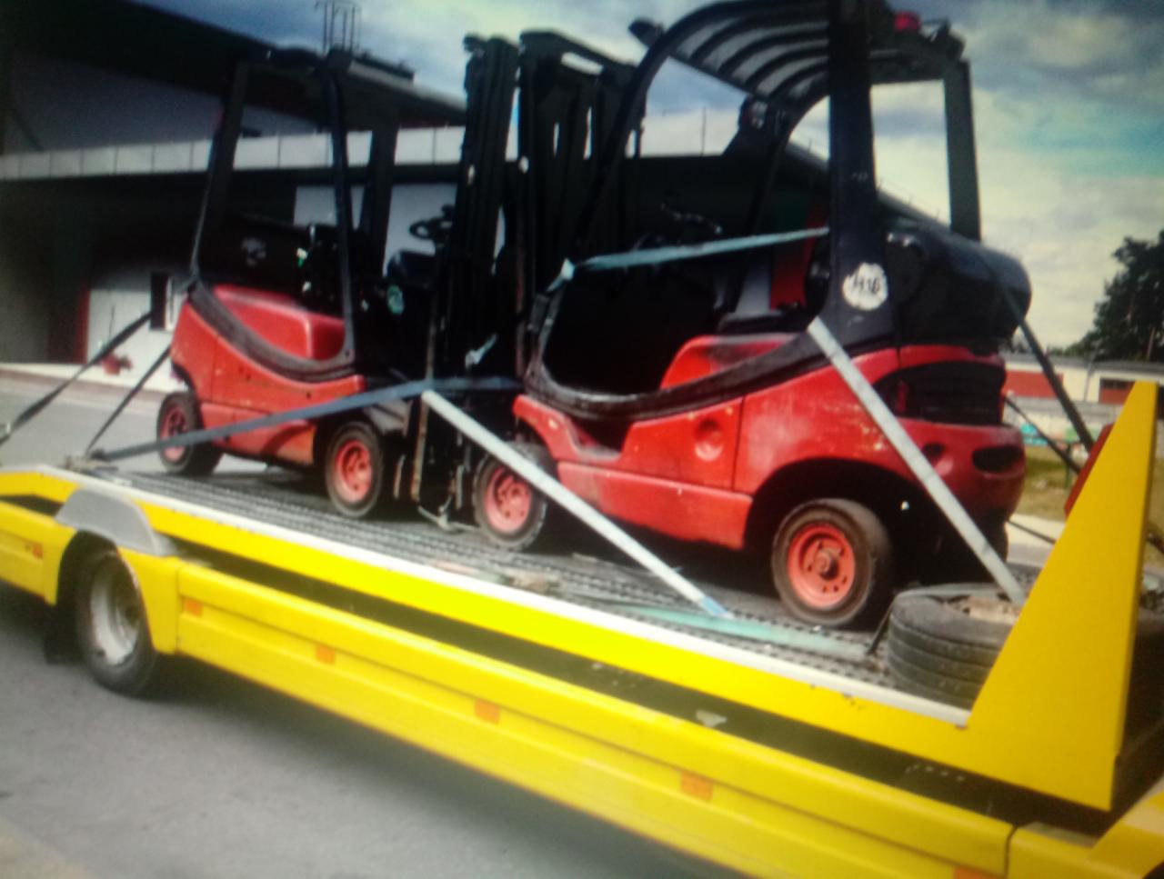 Dwa czerwone wózki widłowe, zabezpieczone pasami, przewożone na żółtej platformie lawety. Widoczne ślady użytkowania na wózkach.