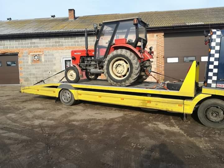 Czerwony traktor Ursus na żółtej platformie lawety, zabezpieczony pasami transportowymi, na tle budynku z cegły.