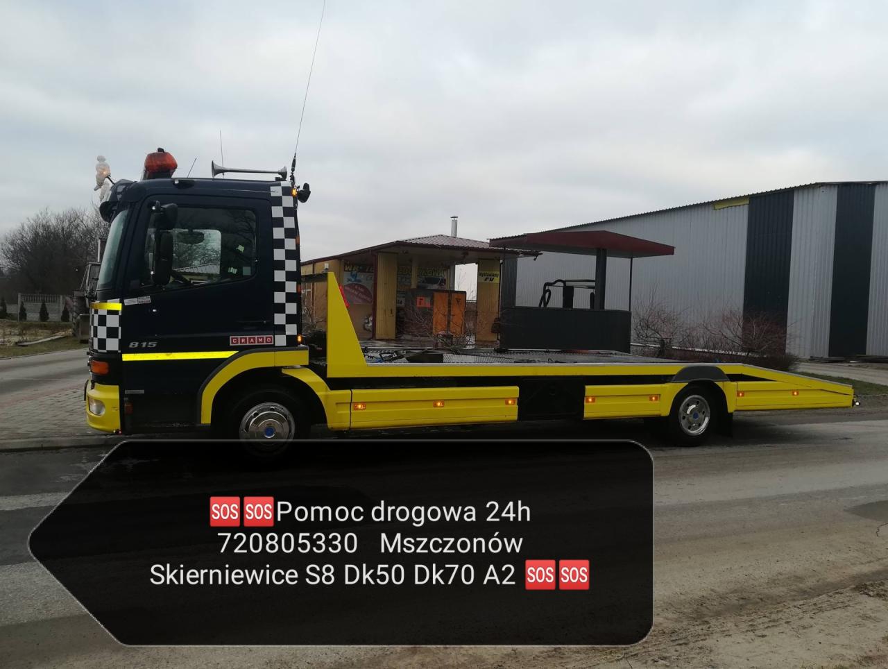 Żółto-czarna laweta Mercedes-Benz z szachownicą na kabinie, zaparkowana na szarej drodze, widoczny budynek w tle i naklejka z numerem telefonu pomocy drogowej.