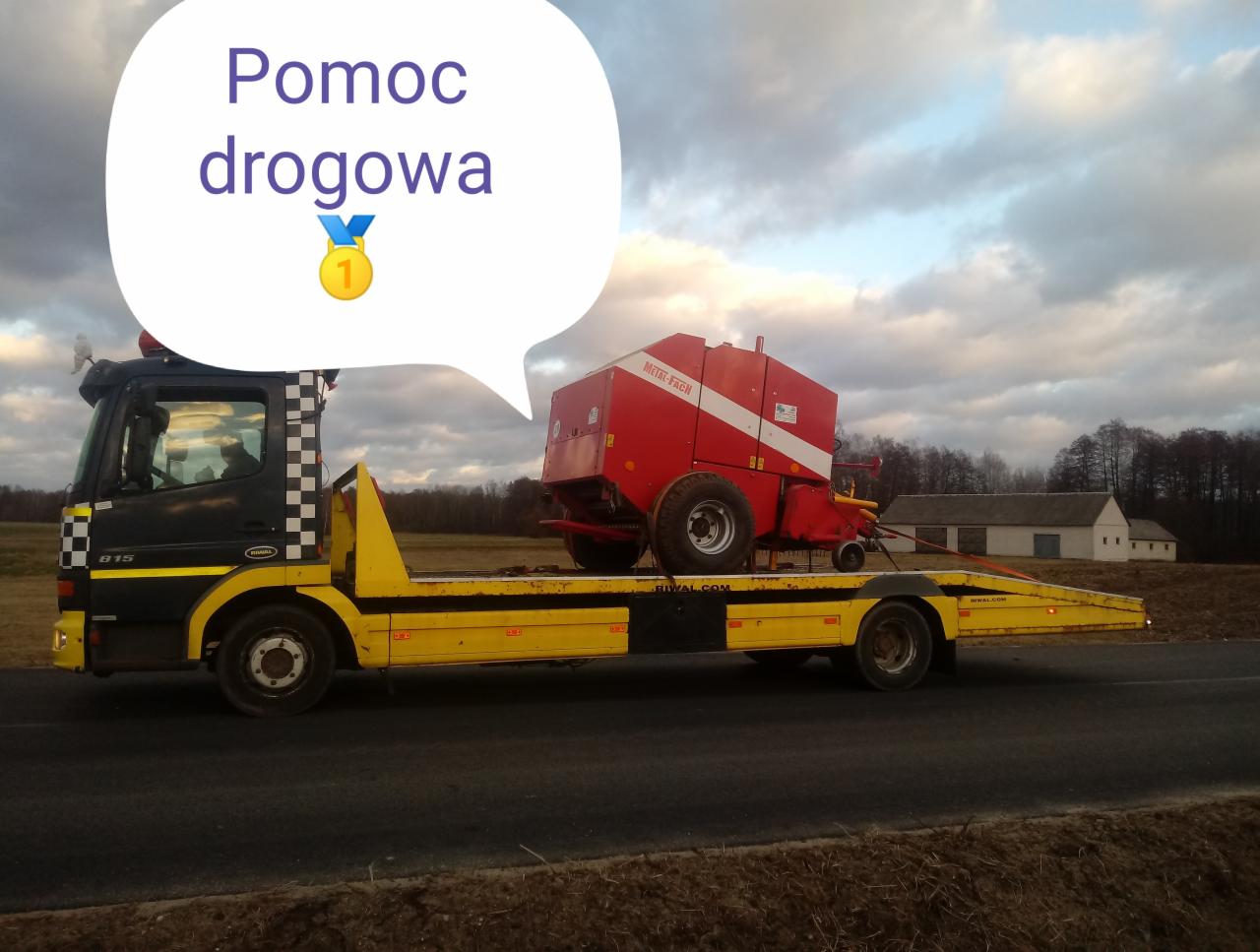 Żółto-czarna laweta przewozi czerwoną prasę rolniczą na tle pochmurnego nieba i zabudowań gospodarczych. Na zdjęciu widoczny dymek z napisem 'Pomoc drogowa' i ikoną medalu.