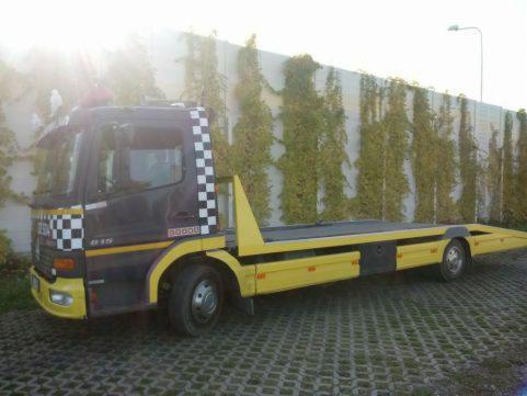 Żółto-czarna laweta z szachownicą na kabinie, stojąca na kostce brukowej przed betonowym murem porośniętym bluszczem.