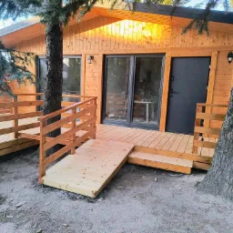 Drewniany dom szkieletowy z tarasem i rampą dla wózków inwalidzkich, otoczony drzewami.