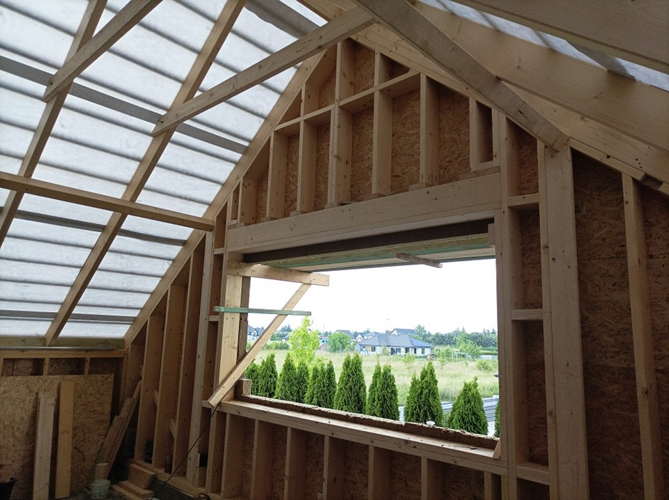 Wnętrze domu szkieletowego w trakcie budowy, widoczna drewniana konstrukcja dachu i ścian z otworem okiennym z widokiem na zielone otoczenie.