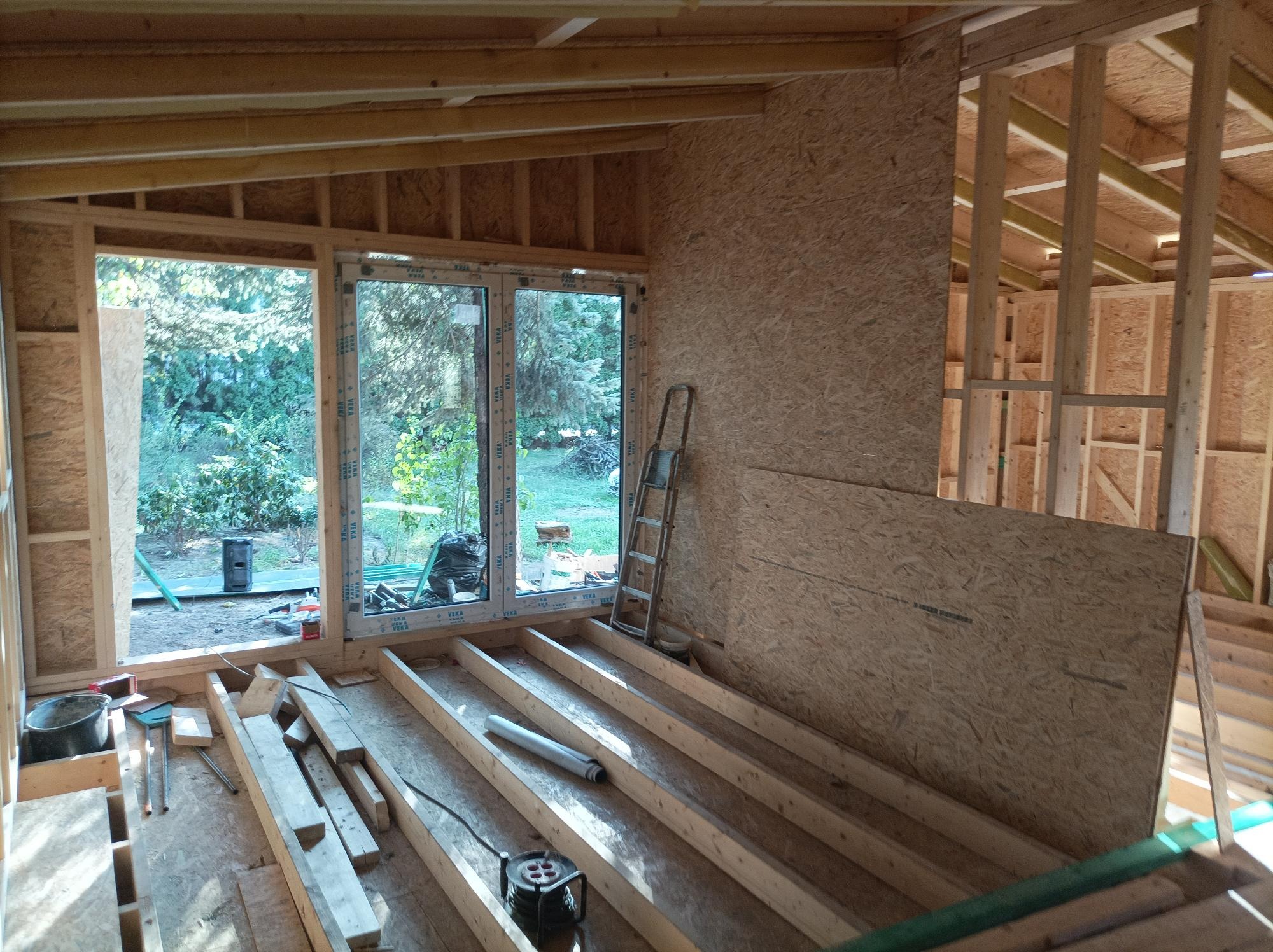 Wnętrze w trakcie budowy: widoczne drewniane elementy konstrukcyjne, okno z widokiem na zieleń, drabina oparta o ścianę z płyty OSB, rozłożone belki podłogowe i narzędzia budowlane.