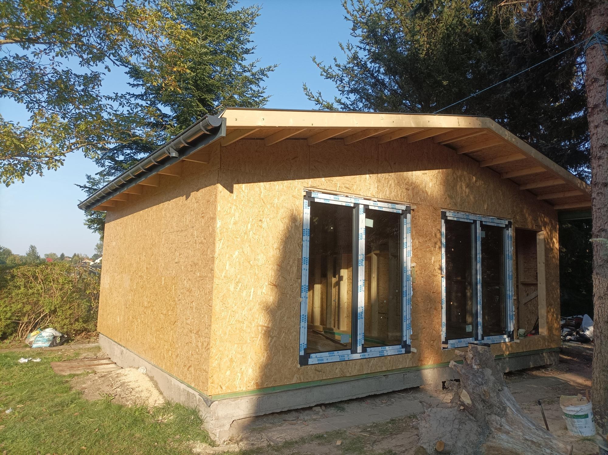 Surowy stan domu szkieletowego z płyt OSB, widoczne okna z folią ochronną i przygotowane miejsce na kolejne okno, w tle drzewa.