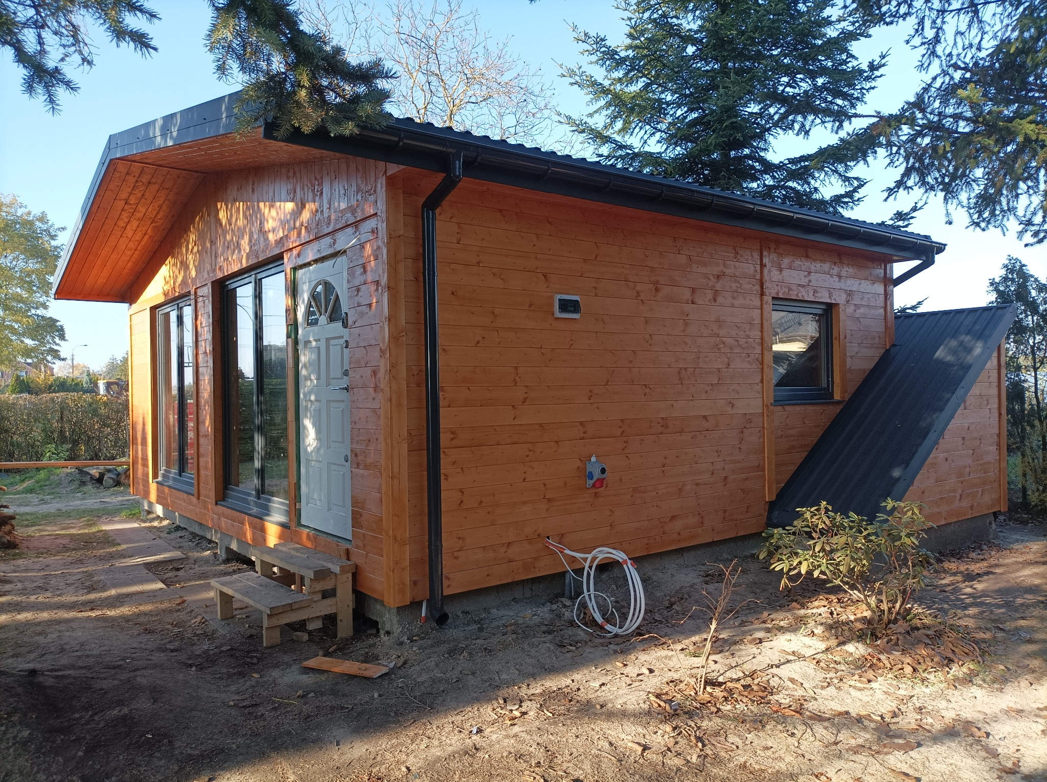Gotowy, drewniany budynek z jasnego drewna z czarnym dachem i rynnami, widoczne schodki wejściowe i niezagospodarowany teren wokół.