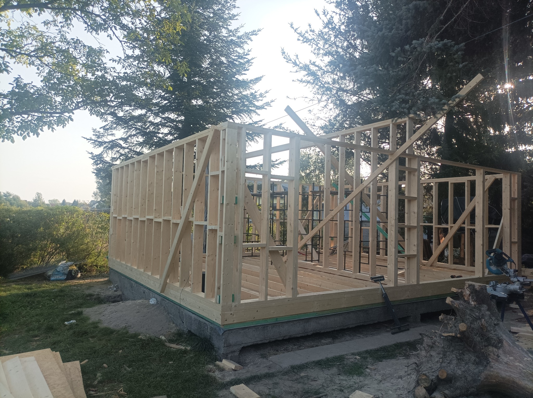 Surowa konstrukcja domu z drewnianych belek na betonowym fundamencie, widoczne narzędzia budowlane i drzewa w tle.