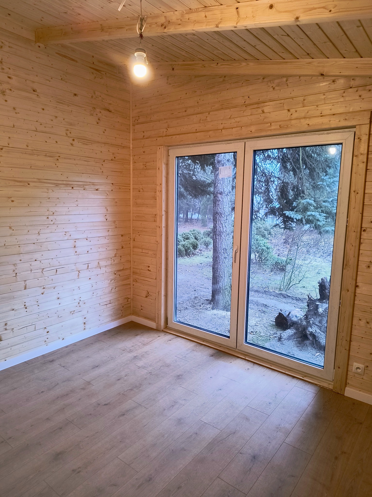 Wnętrze wykończonego pokoju w domu z drewnianą boazerią i podłogą, z widokiem na zewnątrz przez duże okno tarasowe.