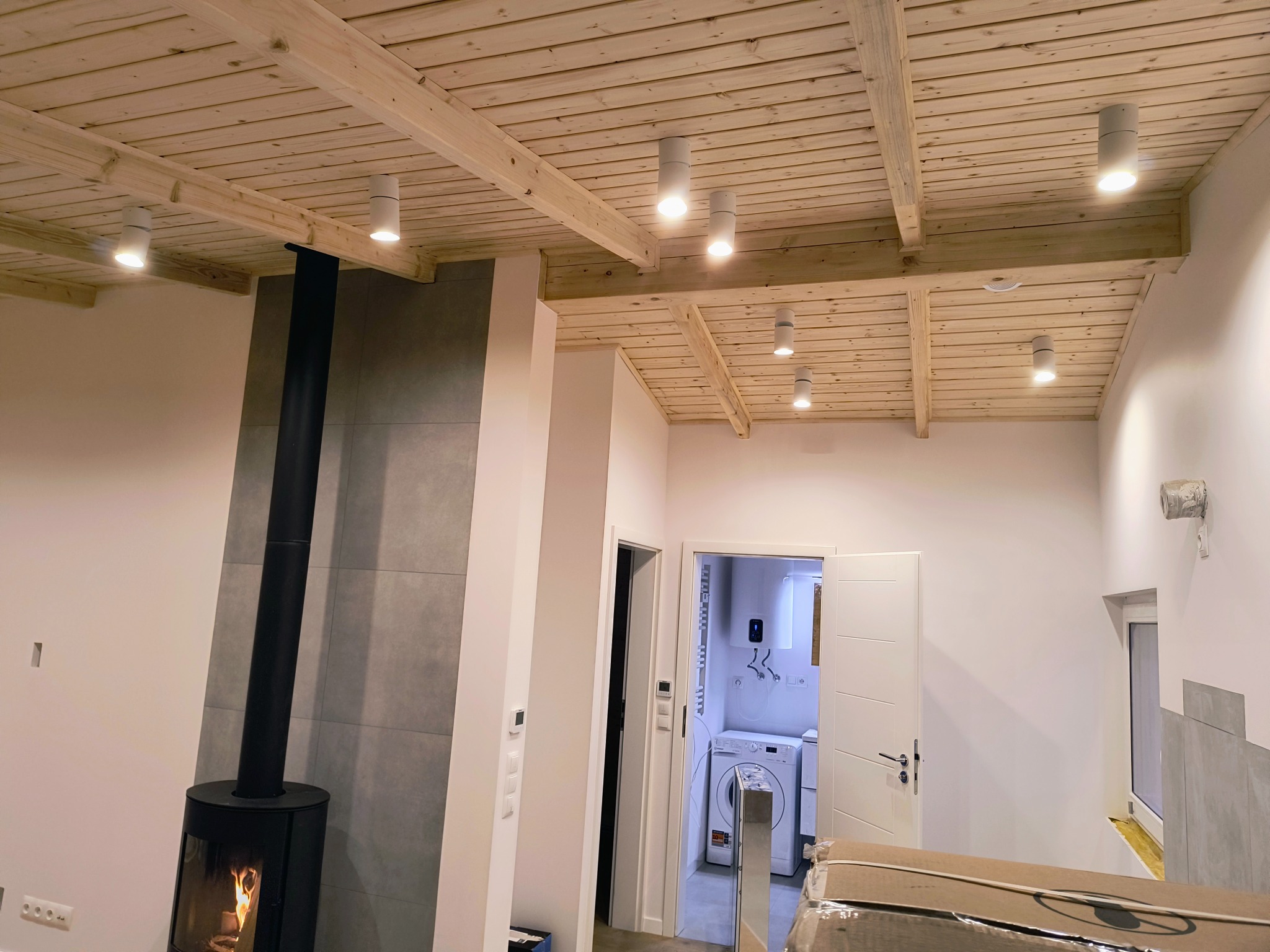 Wnętrze domu w trakcie wykończenia: drewniany sufit z belkami i punktowym oświetleniem, kominek z czarną rurą, łazienka z pralką widoczna w tle.