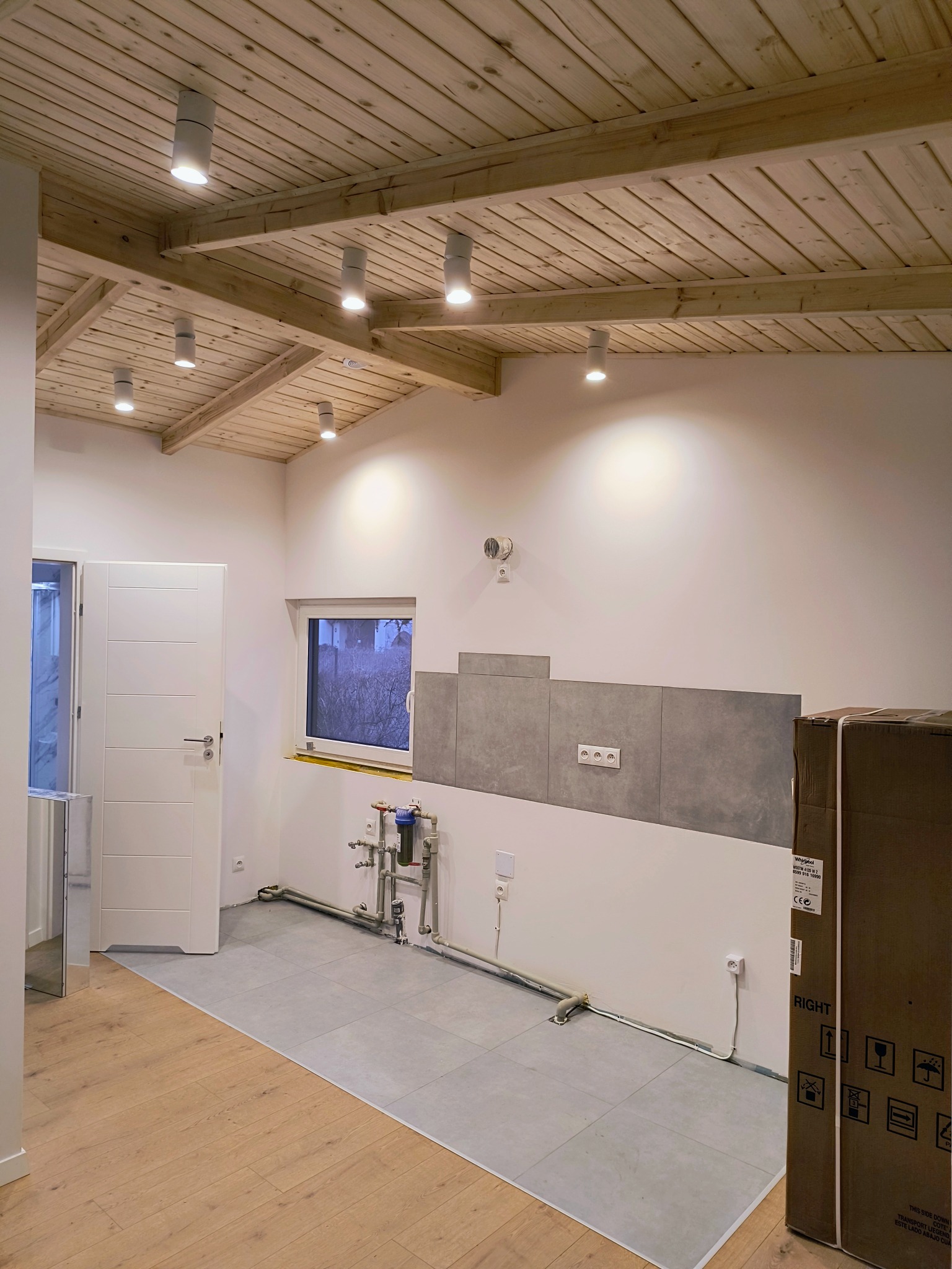 Wnętrze domu w stanie deweloperskim z drewnianym sufitem z oświetleniem punktowym, szare płytki na podłodze i ścianie, instalacja wodna, białe drzwi i kartonowe pudło w rogu pokoju.