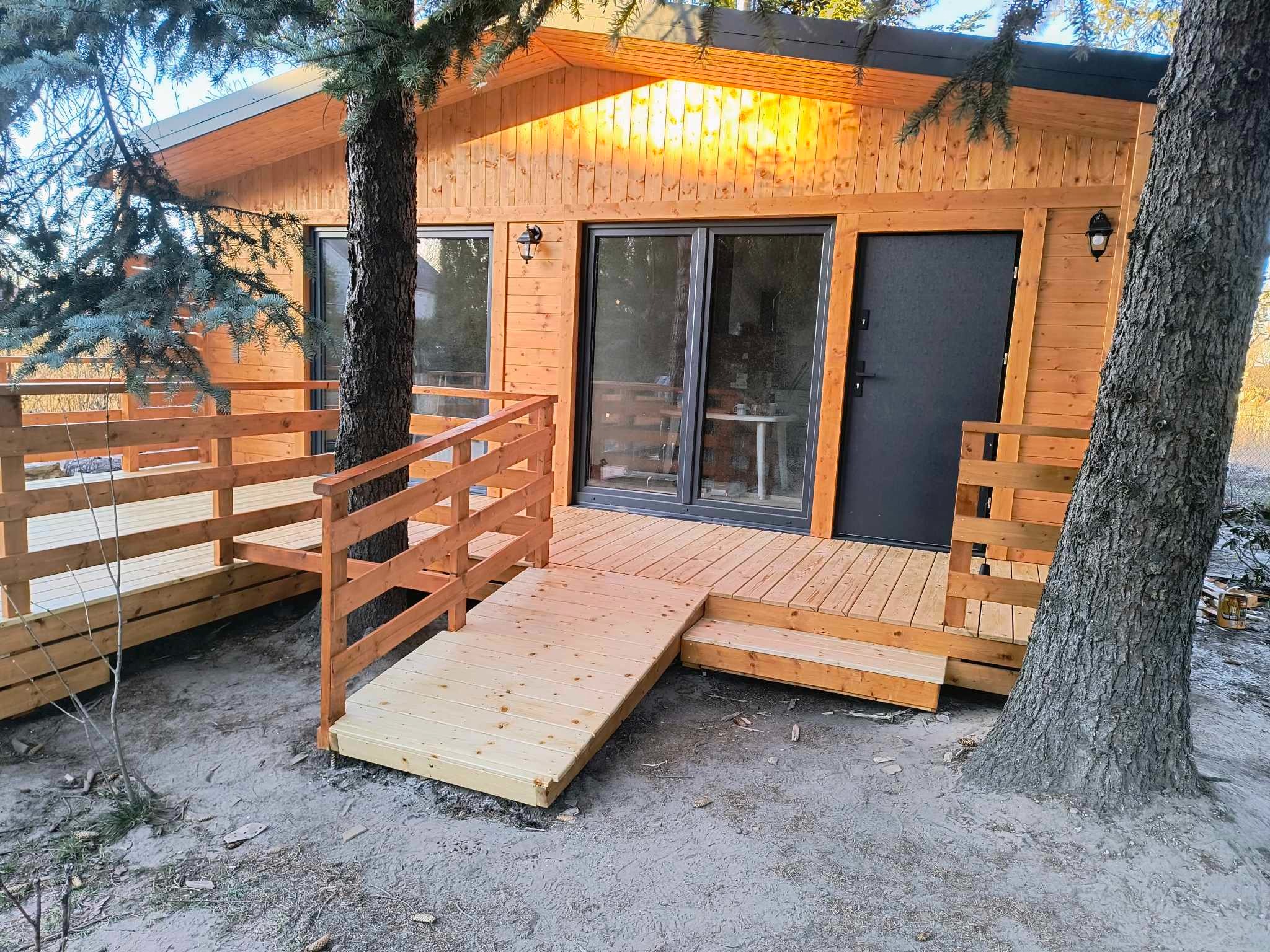 Drewniany dom szkieletowy z tarasem i rampą dla wózków inwalidzkich, otoczony drzewami.