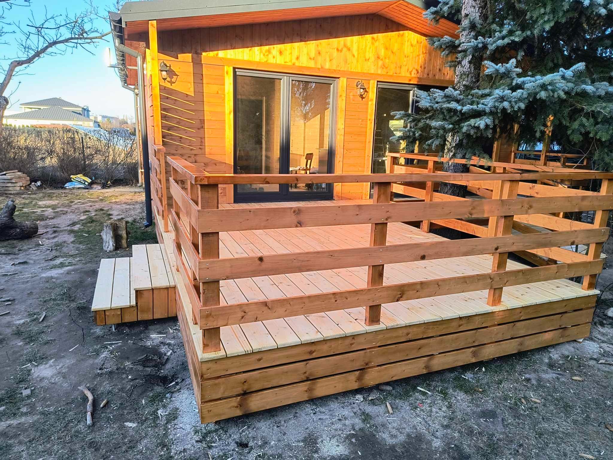 Nowo wybudowany drewniany taras z balustradą przy domku, widoczne schodki wejściowe, błękitny świerk w tle.