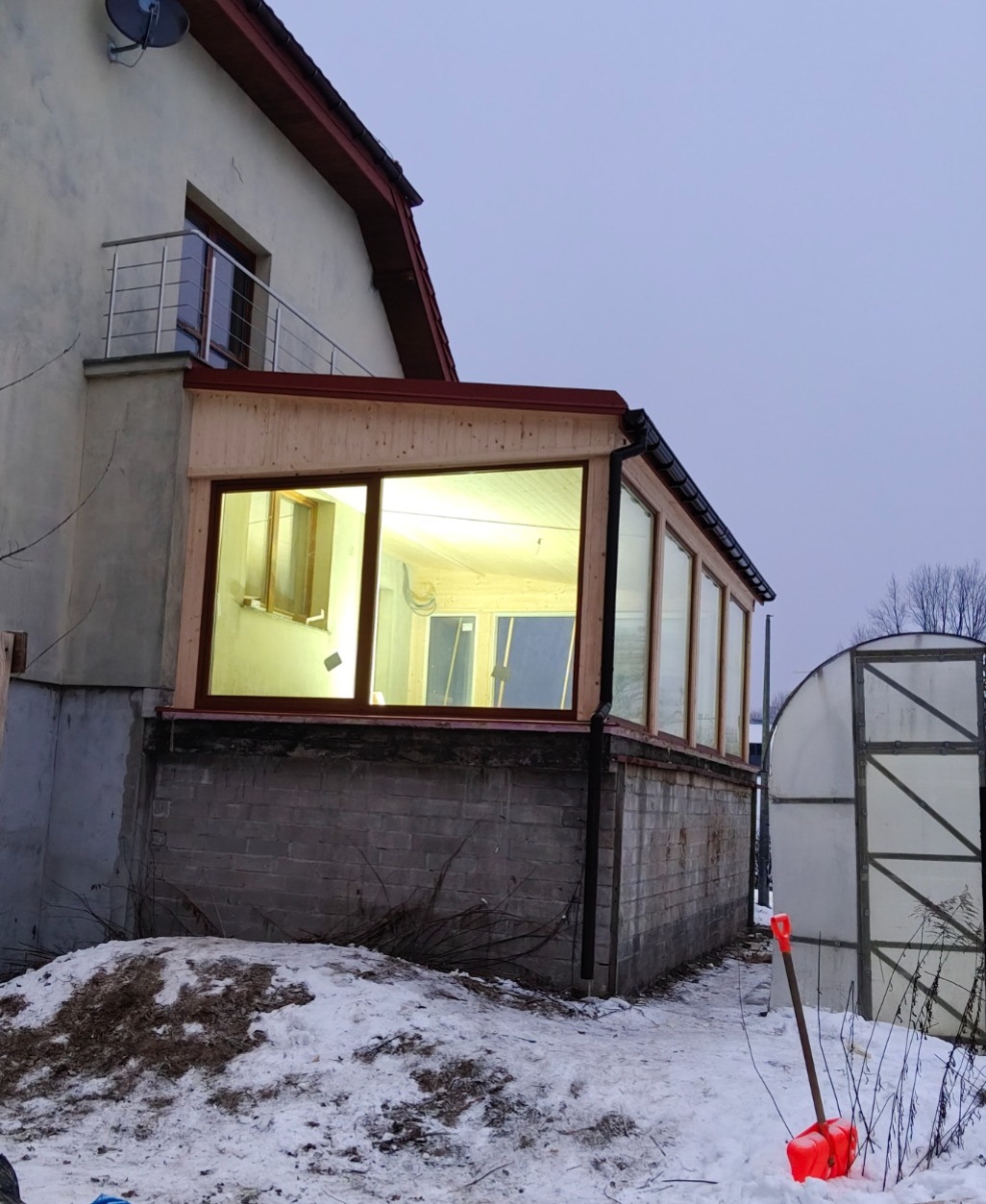 Dobudówka do domu z dużymi oknami, widok z zewnątrz w zimowej scenerii. Czerwona łopata do śniegu oparta o szklarnię w tle.