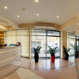 Fotografia Mariusz Olszowski - Panoramiczne ujęcie lobby hotelowego z recepcją oświetloną ledowymi paskami, roślinami doniczkowymi w czarnych i czerwonej osłonce oraz widokiem na ulicę przez szklane drzwi.