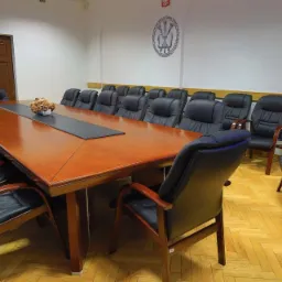 Sala konferencyjna z długim drewnianym stołem, rzędem czarnych skórzanych krzeseł z drewnianymi podłokietnikami, parkietem w jodełkę i godłem na ścianie.