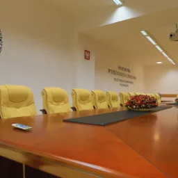 Sala konferencyjna z długim drewnianym stołem, żółtymi skórzanymi krzesłami, logo uczelni na ścianie i kwiatami na stole.