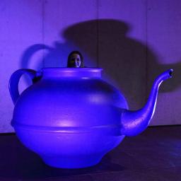 Fotografia Mariusz Olszowski - Kobieta wystająca z wnętrza gigantycznego, niebieskiego czajnika na tle betonowej ściany oświetlonej fioletowym światłem.