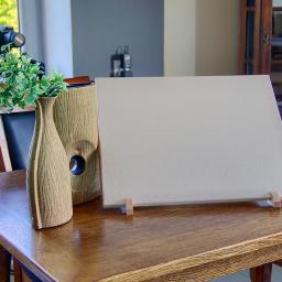 Fotografia Mariusz Olszowski - Beżowa, teksturowana tablica na drewnianym stole, wsparta na małych drewnianych nóżkach, obok wazon z zielonymi gałązkami i drugi wazon z otworem, w tle aparat na statywie i okno.