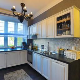 Fotografia Mariusz Olszowski - Jasna kuchnia z białymi szafkami w stylu klasycznym, czarnym blatem, mozaiką na ścianie, oknem z roletami rzymskimi i czarnym żyrandolem.