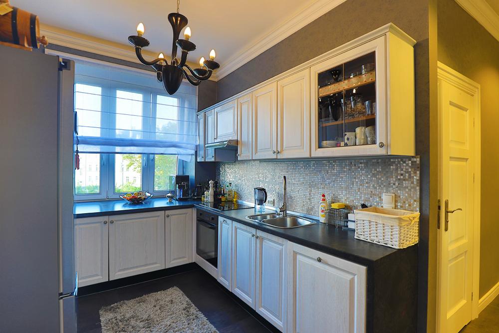 Jasna kuchnia z białymi szafkami w stylu klasycznym, czarnym blatem, mozaiką na ścianie, oknem z roletami rzymskimi i czarnym żyrandolem.