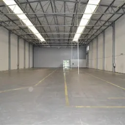 Pusta hala magazynowa w Warszawie z betonową podłogą oznaczona żółtymi liniami, widoczne stalowe słupy konstrukcyjne i świetliki w dachu.