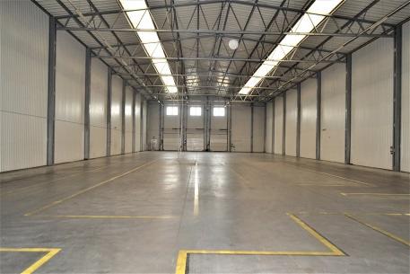 Pusty magazyn z betonową podłogą i żółtymi liniami wyznaczającymi strefy, widoczne stalowe belki konstrukcyjne i białe ściany z panelami.