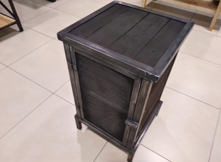 Metalowa szafka nocna z drewnianym blatem i siatką w industrialnym stylu, stojąca na jasnych płytkach.