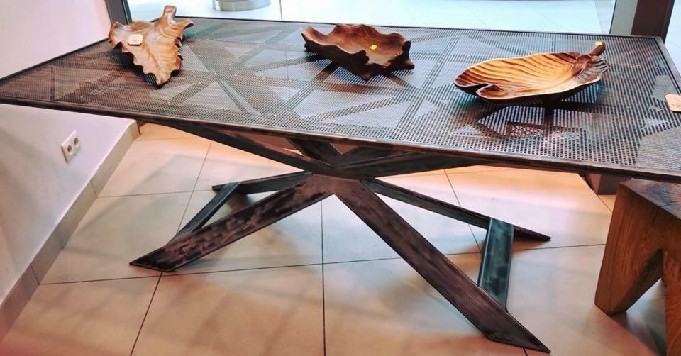 Stół z metalowym blatem o siatkowej strukturze i industrialnej podstawie w kształcie gwiazdy, na blacie trzy drewniane misy w kształcie liści, widoczne gniazdko elektryczne w ścianie i fragment...