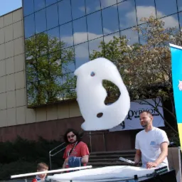 Duża bańka mydlana w kształcie ducha unosząca się w powietrzu podczas pokazu na zewnątrz, z widocznymi osobami i kolorowym banerem z postaciami z kreskówek w tle.