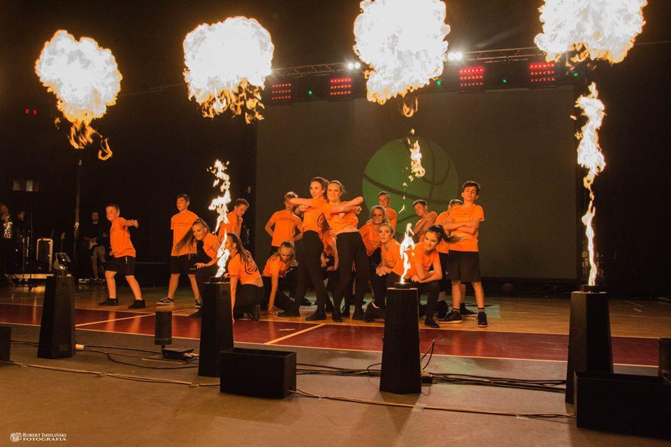 Grupa młodych ludzi w pomarańczowych koszulkach na scenie z efektami pirotechnicznymi; widoczne płomienie ognia oraz dym w tle, na podłodze czarne prostopadłościany