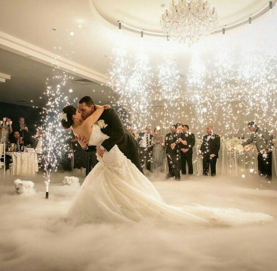 Pocałunek pary młodej otoczonej fontannami iskier i gęstą mgłą na parkiecie podczas wesela w eleganckiej sali balowej z żyrandolem.