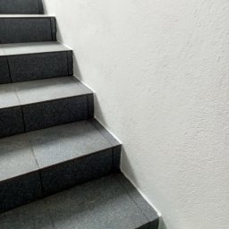 Mirosław Brzózka - Schody wyłożone szarymi, kwadratowymi płytkami ceramicznymi z ciemnoszarymi brzegami, przylegające do białej, teksturowanej ściany.