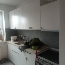 Mirosław Brzózka - Wnętrze kuchni w trakcie remontu: białe szafki z czarnymi uchwytami, szary panel nad blatem, widoczne wiadro z farbą i narzędzia malarskie, niedokończony montaż.