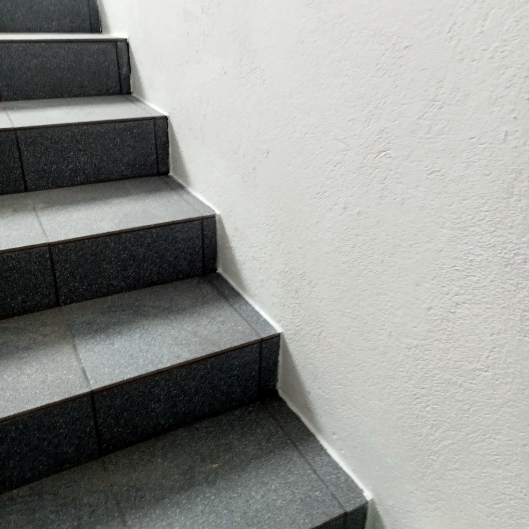Schody wyłożone szarymi, kwadratowymi płytkami ceramicznymi z ciemnoszarymi brzegami, przylegające do białej, teksturowanej ściany.