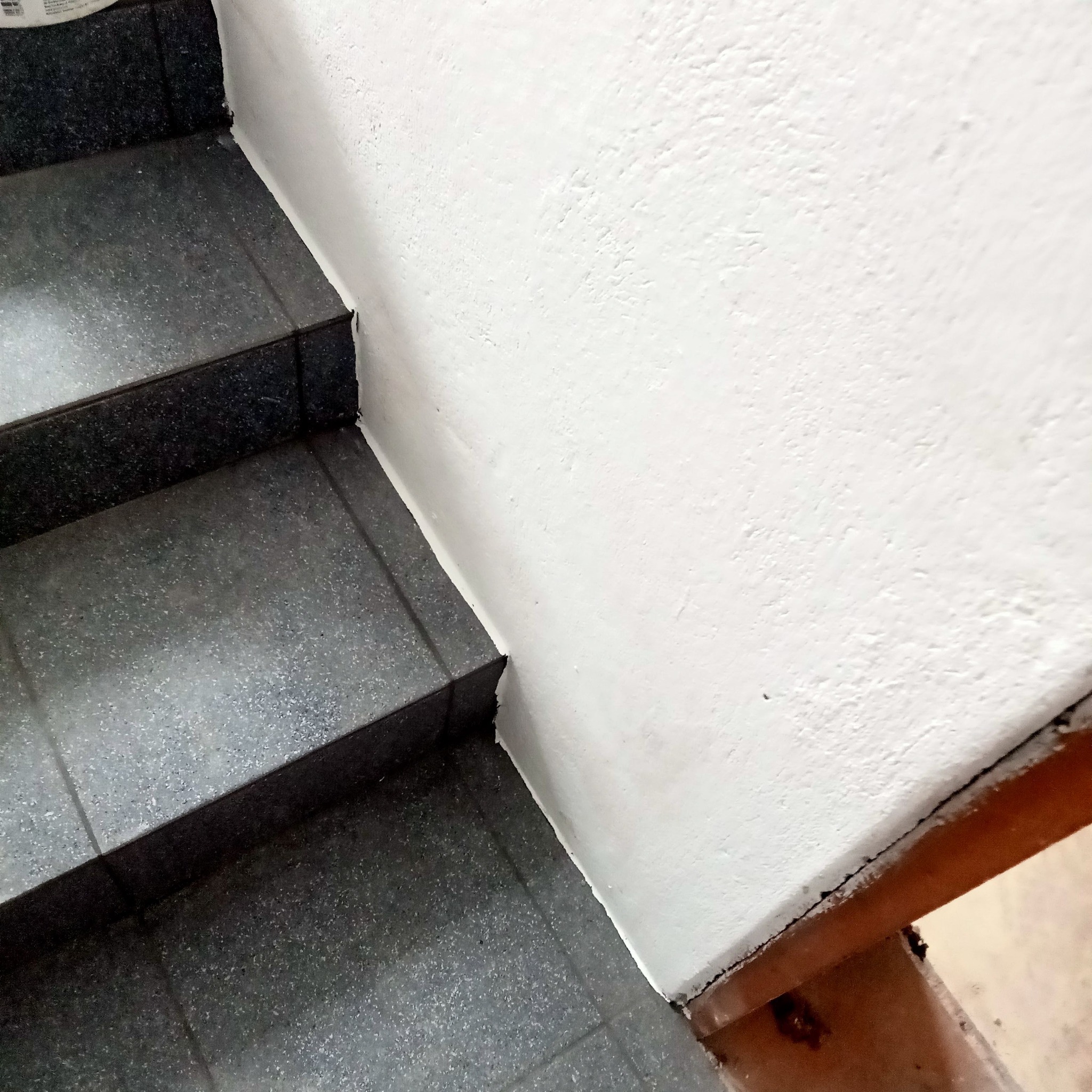 Schody wyłożone ciemnoszarą, błyszczącą glazurą, przylegające do świeżo pomalowanej na biało ściany z widoczną nierównością na styku z podłogą.