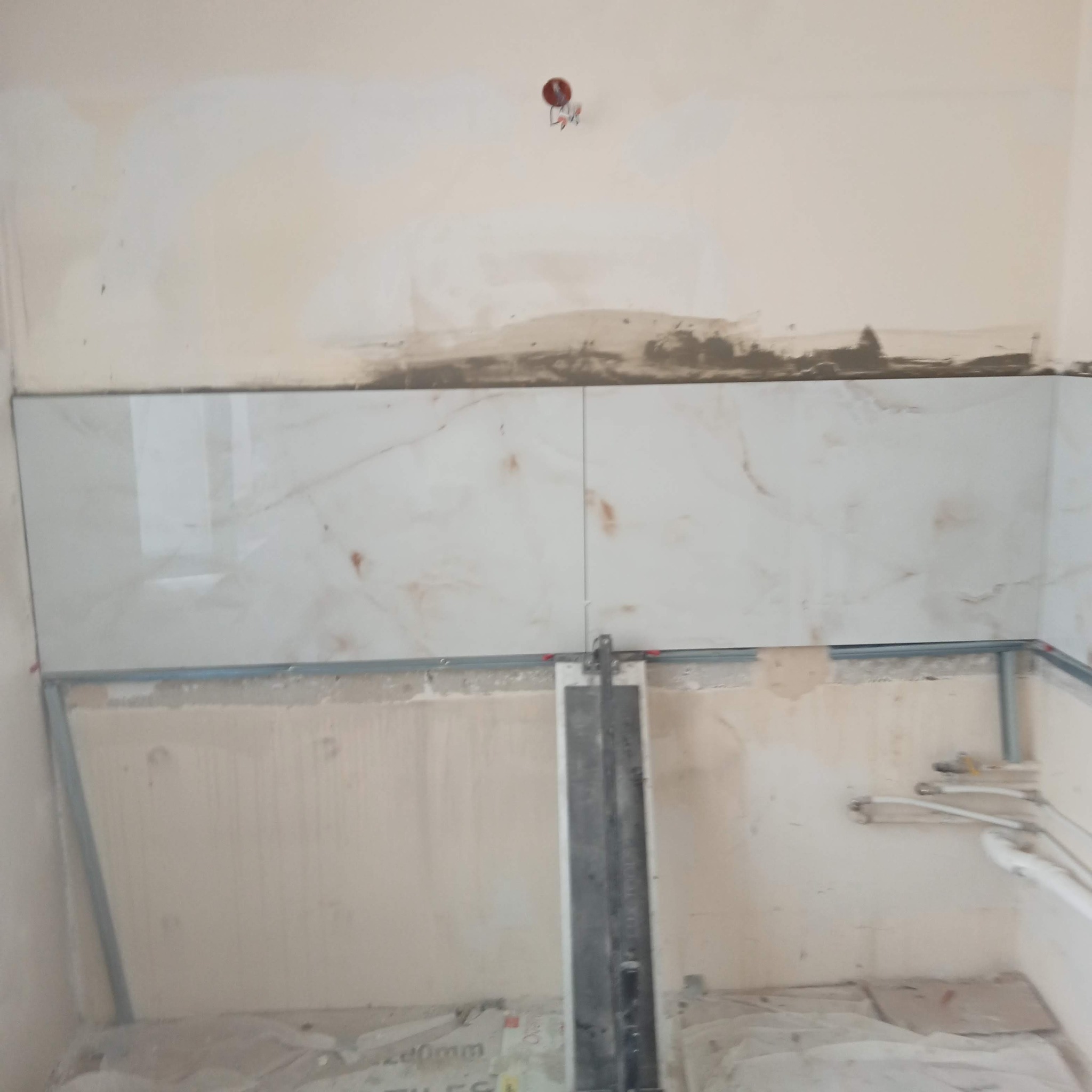 Fragment ściany z ułożonym rzędem jasnych płytek imitujących marmur, widoczne metalowe profile podtrzymujące, ściany w stanie surowym, narzędzie do cięcia płytek na podłodze.