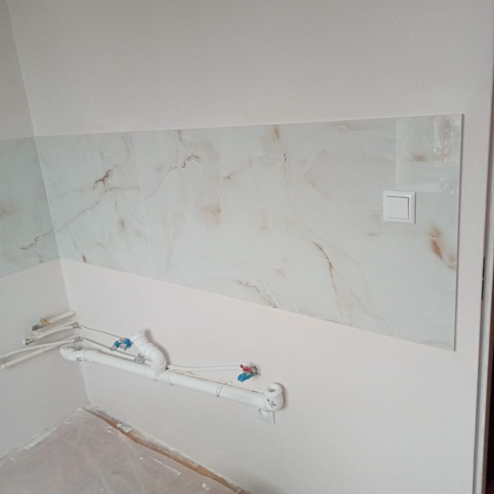Fragment łazienki w trakcie prac: białe płytki imitujące marmur do połowy ściany, widoczne rury i instalacja wodna, surowy stan podłogi zabezpieczonej folią.