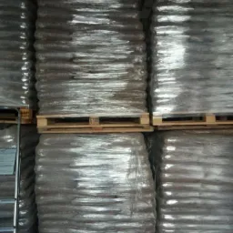 Ułożone na paletach, owinięte folią, brązowe worki z pelletem w magazynie z widocznym fragmentem ściany z cegieł.