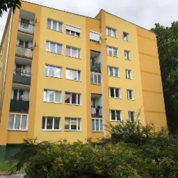 Odnowiony blok mieszkalny w odcieniach żółci z widocznymi balkonami, zieleń krzewów na pierwszym planie i satelita na elewacji.