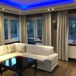 Elegancki salon z narożną sofą w odcieniach bieli, ciemnym stolikiem kawowym z bukietem czerwonych róż, zasłonami w kolorze ecru i oknami z widokiem na zieleń, oświetlony niebieskim światłem LED...