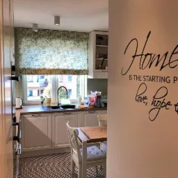 Kuchnia z białymi szafkami, wzorzystą podłogą i oknem przesłoniętym materiałem w roślinny wzór, widok zza narożnika ściany z napisem 'Home is the starting point of love, hope & dreams'.