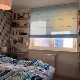 Jasna, błękitna tkanina okienna w stylu rzymskim, zamontowana w sypialni z półkami na książki po bokach okna.