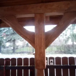 Drewniana konstrukcja dachu altany z widocznym słupem i belkami, na tle ogrodzenia i drzew w zimowej scenerii.