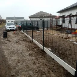 Częściowo zamontowane, czarne ogrodzenie panelowe z betonową podmurówką na błotnistym terenie budowy; w tle domy w trakcie budowy i zaparkowane samochody.