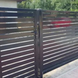 Nowoczesna, przesuwna brama w kolorze antracytu z poziomymi lamelami, zamontowana przy ogrodzeniu z siatki i betonowej podmurówki, widoczna z boku na tle zieleni.