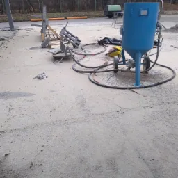 Niebieski, mobilny zbiornik do obróbki strumieniowo-ściernej na betonowej powierzchni placu, obok leżą elementy metalowe na palecie i zwinięte węże.