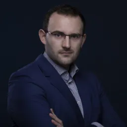 Portret mężczyzny w okularach i niebieskim garniturze, skrzyżowane ręce, ciemne tło.