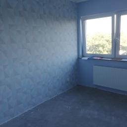 usługi malarskie Marcel - Pomalowane na niebiesko ściany pokoju z geometrycznym wzorem trójkątów, okno z białymi ramami i grzejnik pod nim, surowa podłoga.